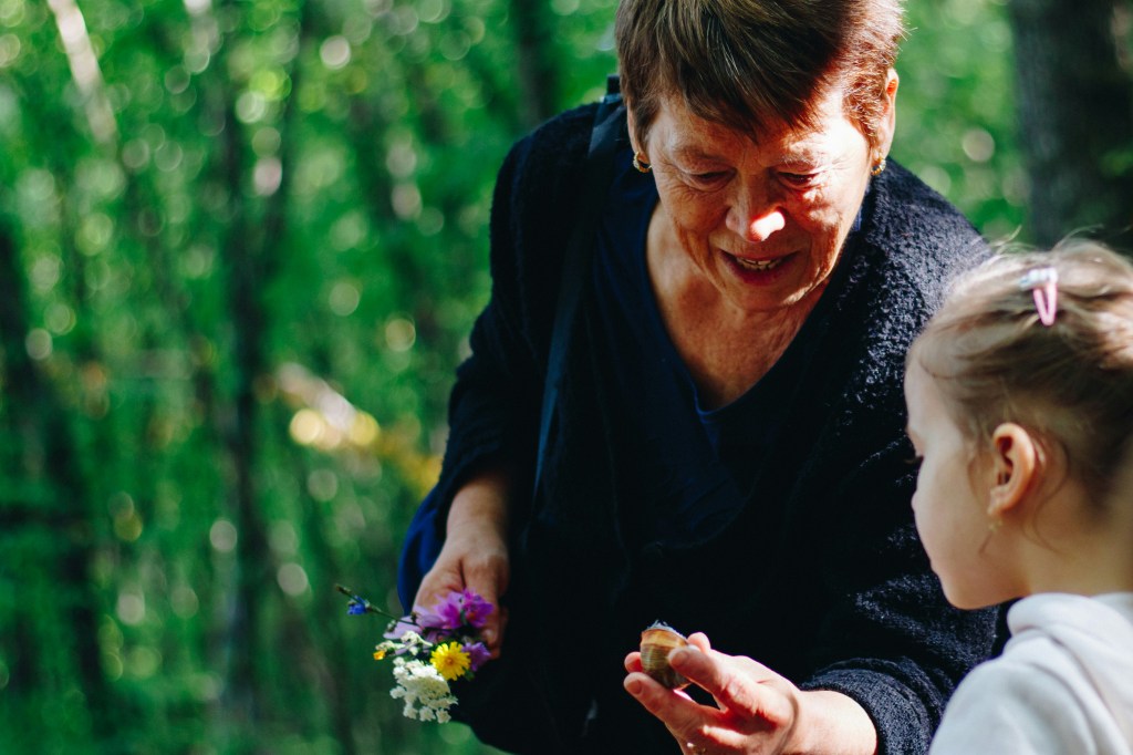 An older woman  showing flowers and a snail to a little girl against a blurry background of green trees