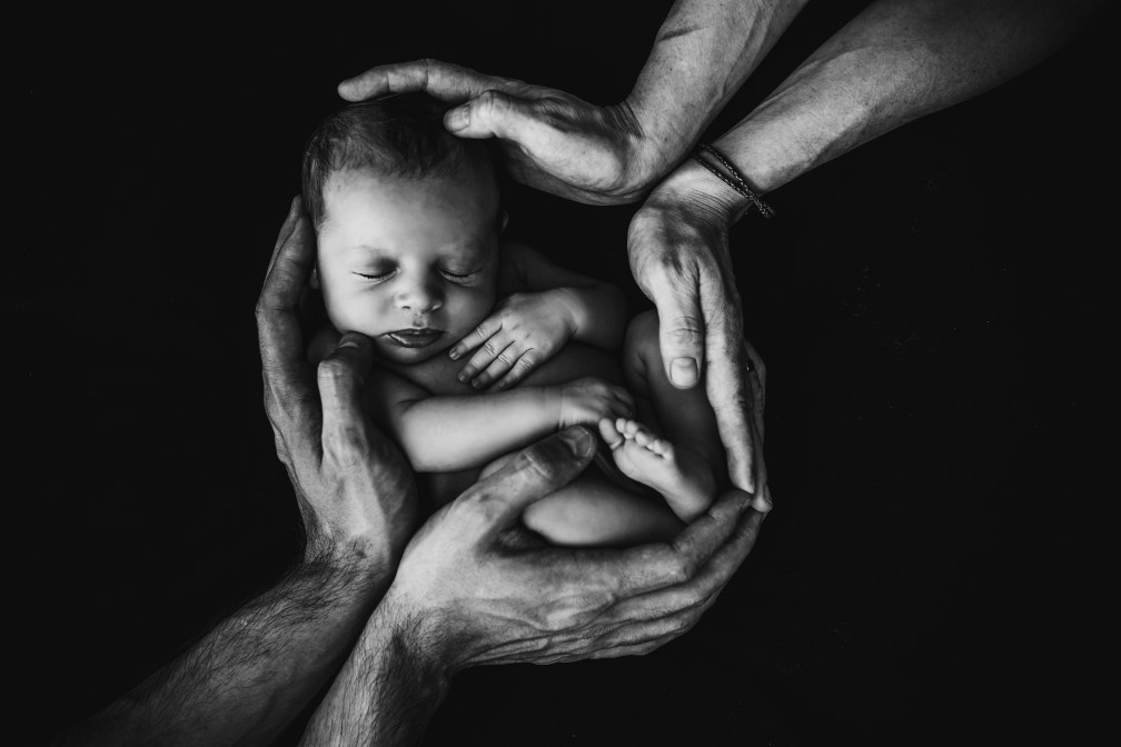 Greyscale photo of a baby being held by two pairs of hands.